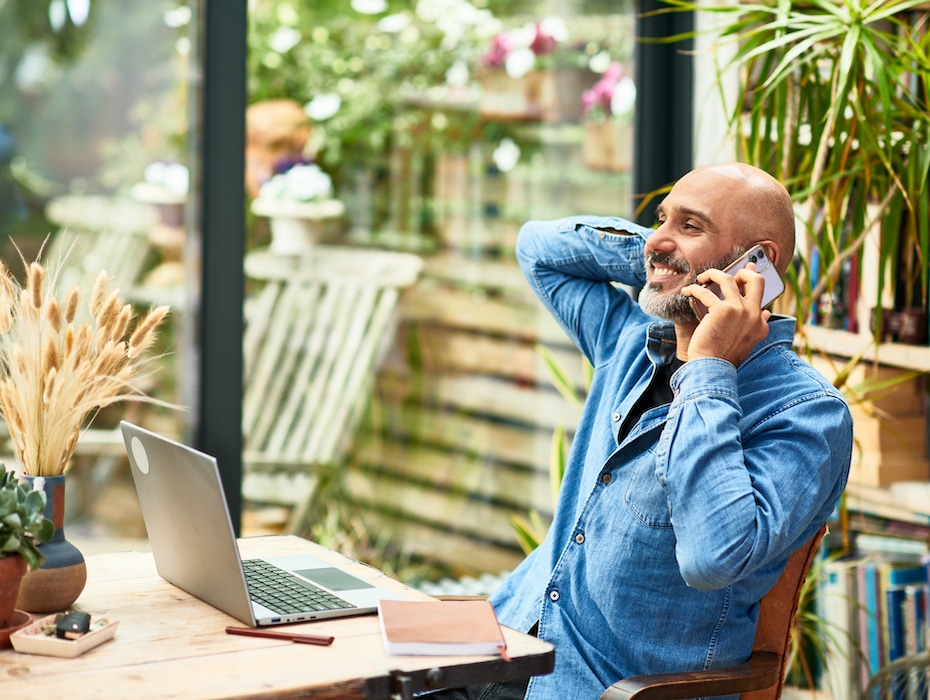 Small business owner leaning back and smiling using mobile phone, working from home, sitting at desk in front of laptop using Microsoft Teams and telephony provided by a UCaaS provider