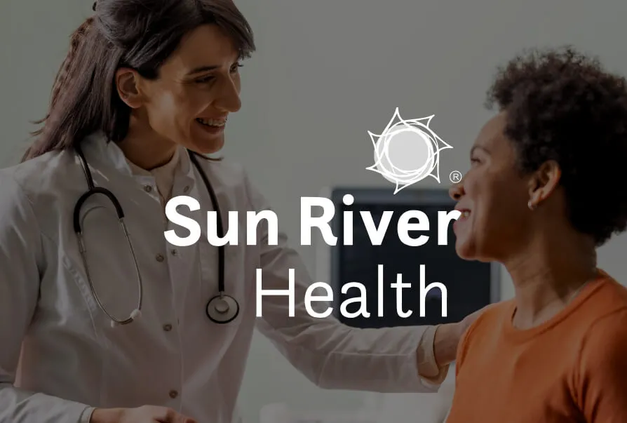 Sun River Health Logo in front of a doctor smiling while touching the shoulder of a patient