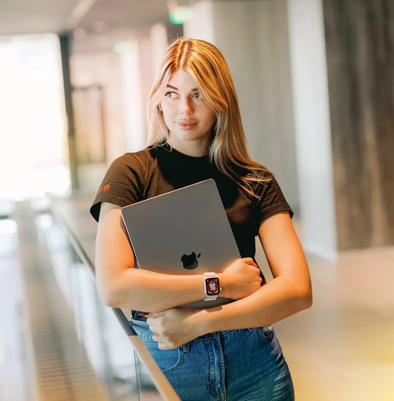 Young woman holding a laptop, wearing a smartwatch, standing in a hallway.