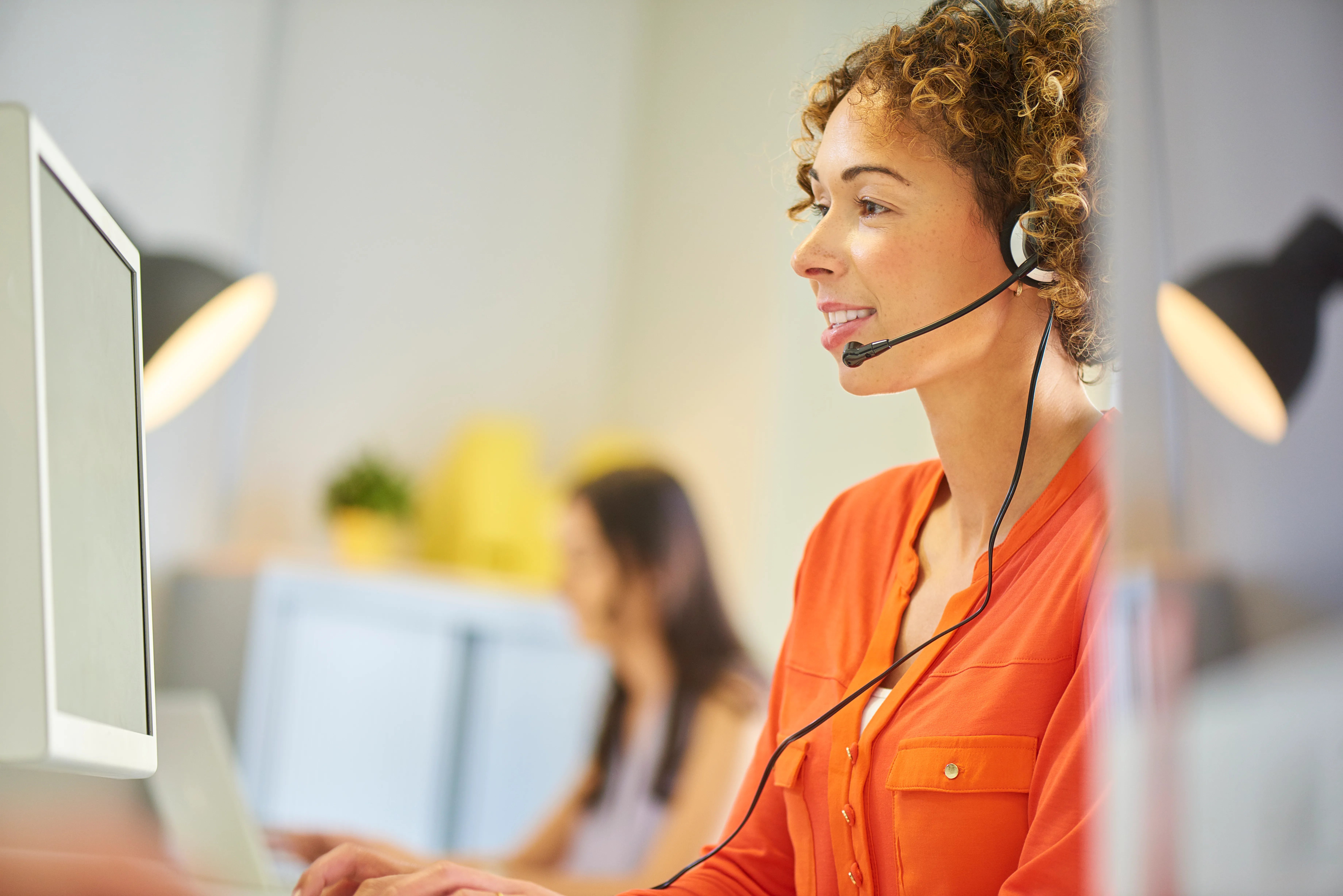 A woman with curly hair wearing a headset and working at a computer in a modern office setting.