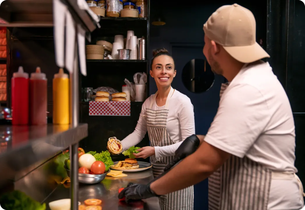 Two kitchen workers in striped aprons smile and talk while assembling burgers on a stainless steel counter filled with fresh ingredients.