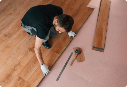 A man installs wood flooring, carefully laying down planks in a well-lit room.
