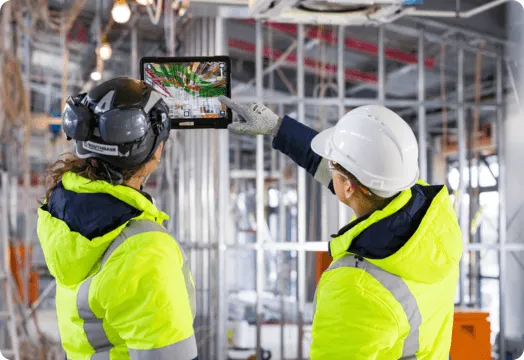 1. Two construction workers in safety vests and hard hats hold a tablet, discussing plans on a job site.