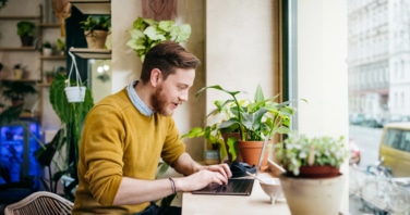 A man smiling while working from home while being surrounded by house plants.
