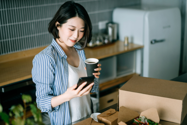 Beautiful smiling young Asian woman standing by the kitchen counter, using smartphone while enjoying assorted home delivery takeaway food, coffee and fresh green salad at home. Eating at home concept-273