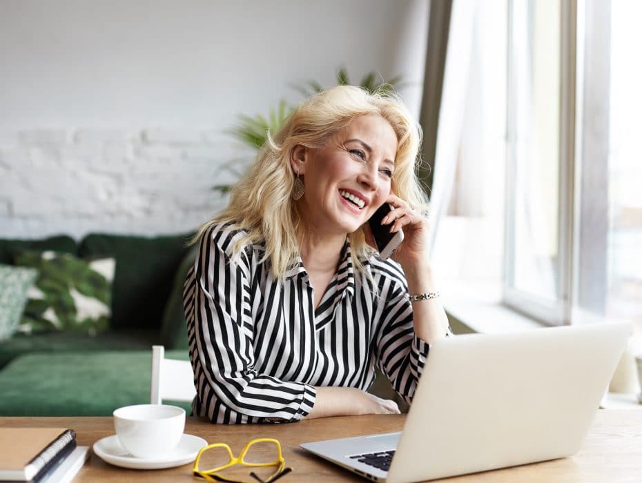 Attractive successful elderly businesswoman in striped blouse working in modern office, making phone call to potential client, having nice conversation, sitting at desk in front of open laptop