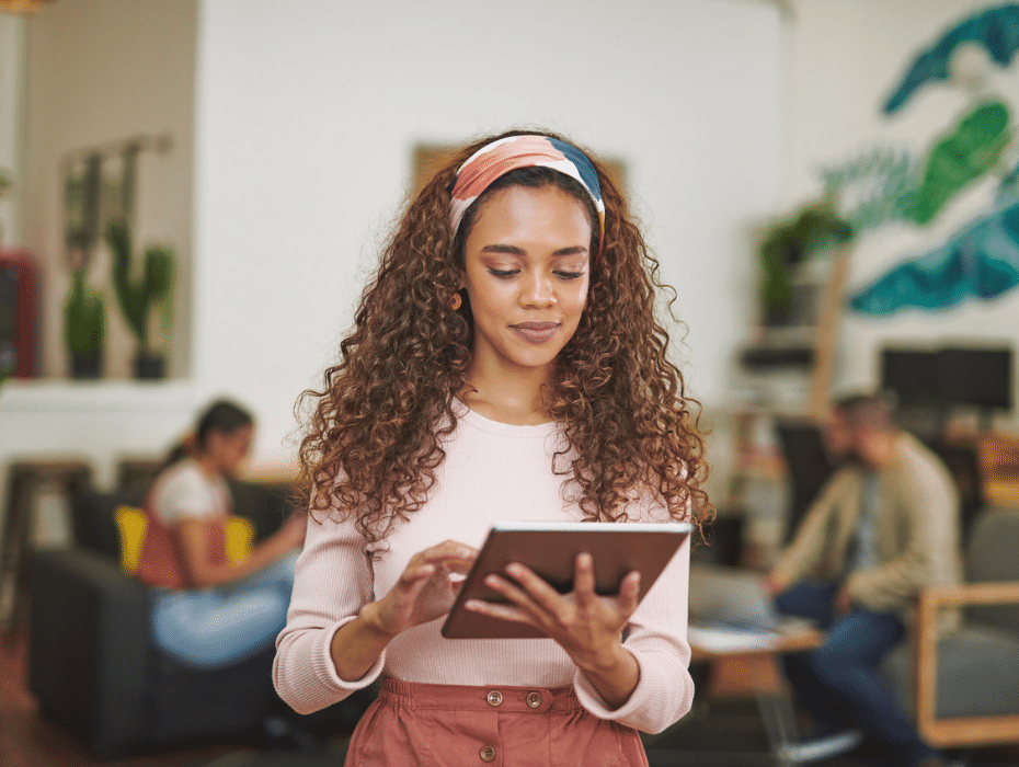 Woman on iPad standing in an office with coworkers chatting behind her.