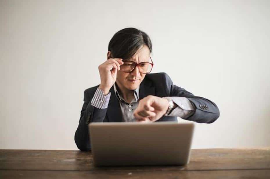 Governmental employee with glasses checking the time on his watch while sitting in front of a laptop computer