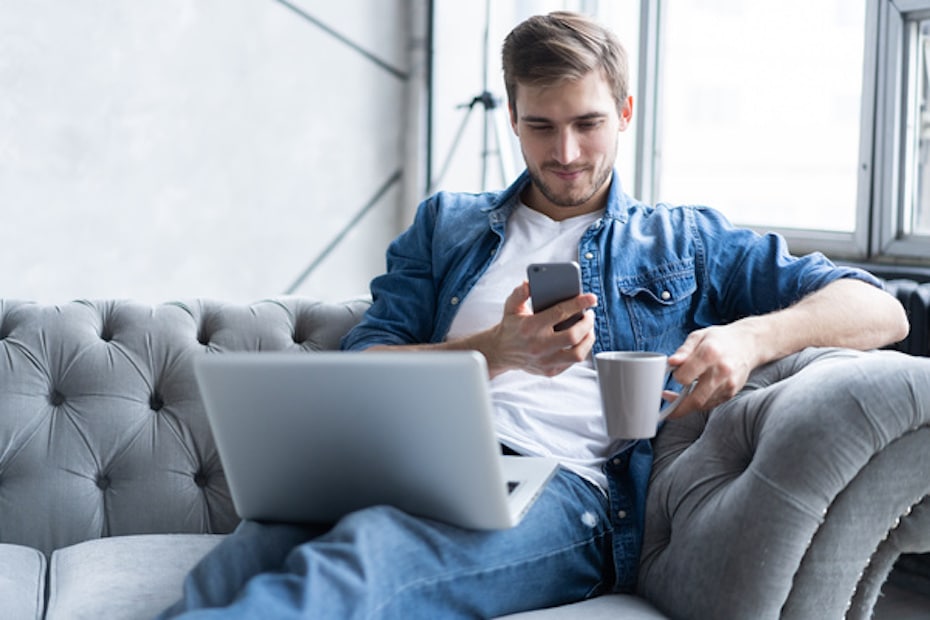 Man sitting on couch with mobile phone and laptop performing online banking as a service