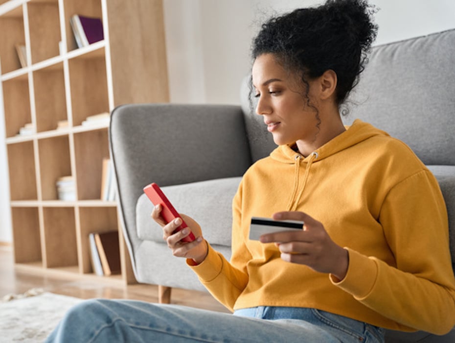 A consumer holds a mobile phone in one hand and a credit card in the other while sitting on the floor leaning against a couch