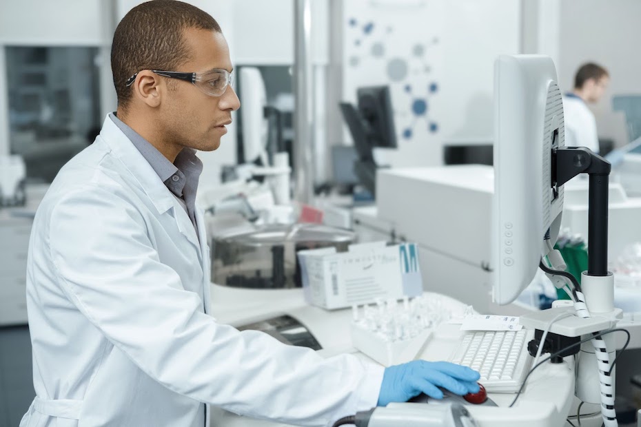 Pharmaceutical company employee wearing protective goggles and gloves stands at a laboratory-style table viewing intellectual property on a computer.