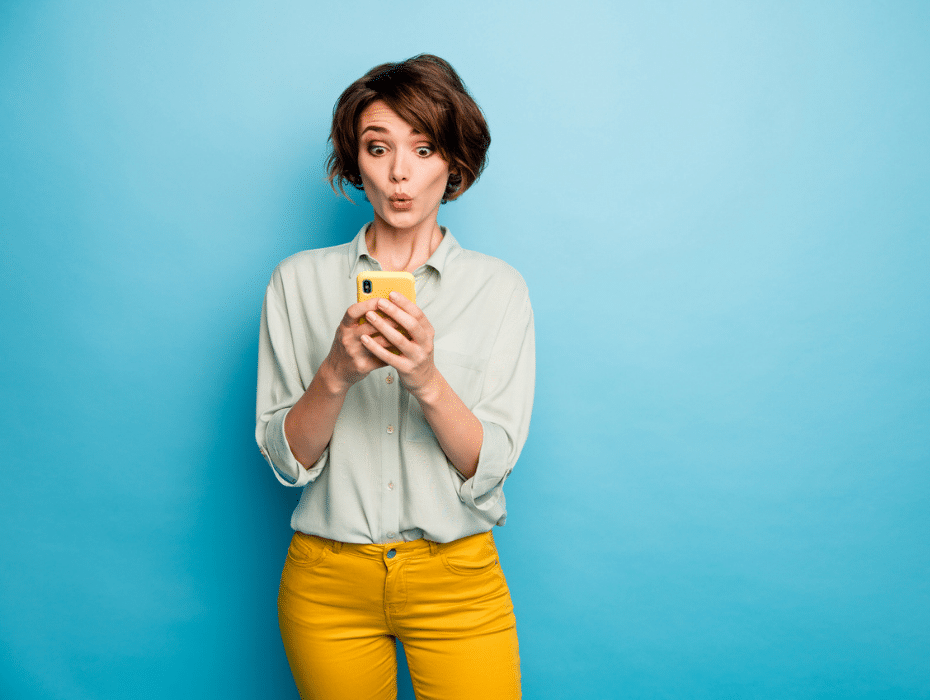 Woman in light blue top and orange pants on a blue background looking pleasantly surprised at her smartphone