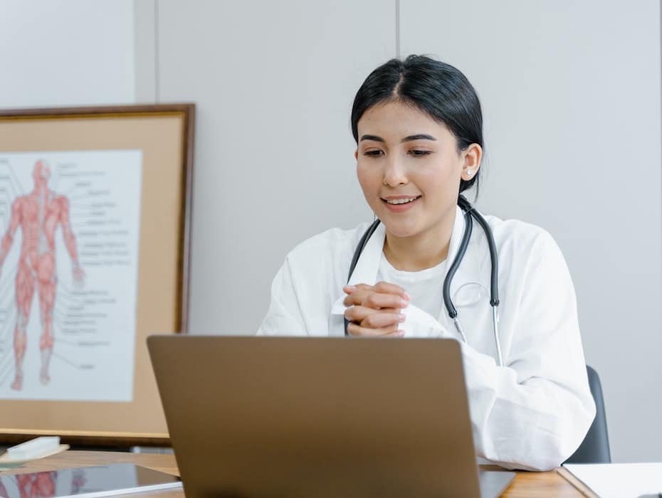 A healthcare provider sits at her desk in front of a laptop for a telehealth appointment.