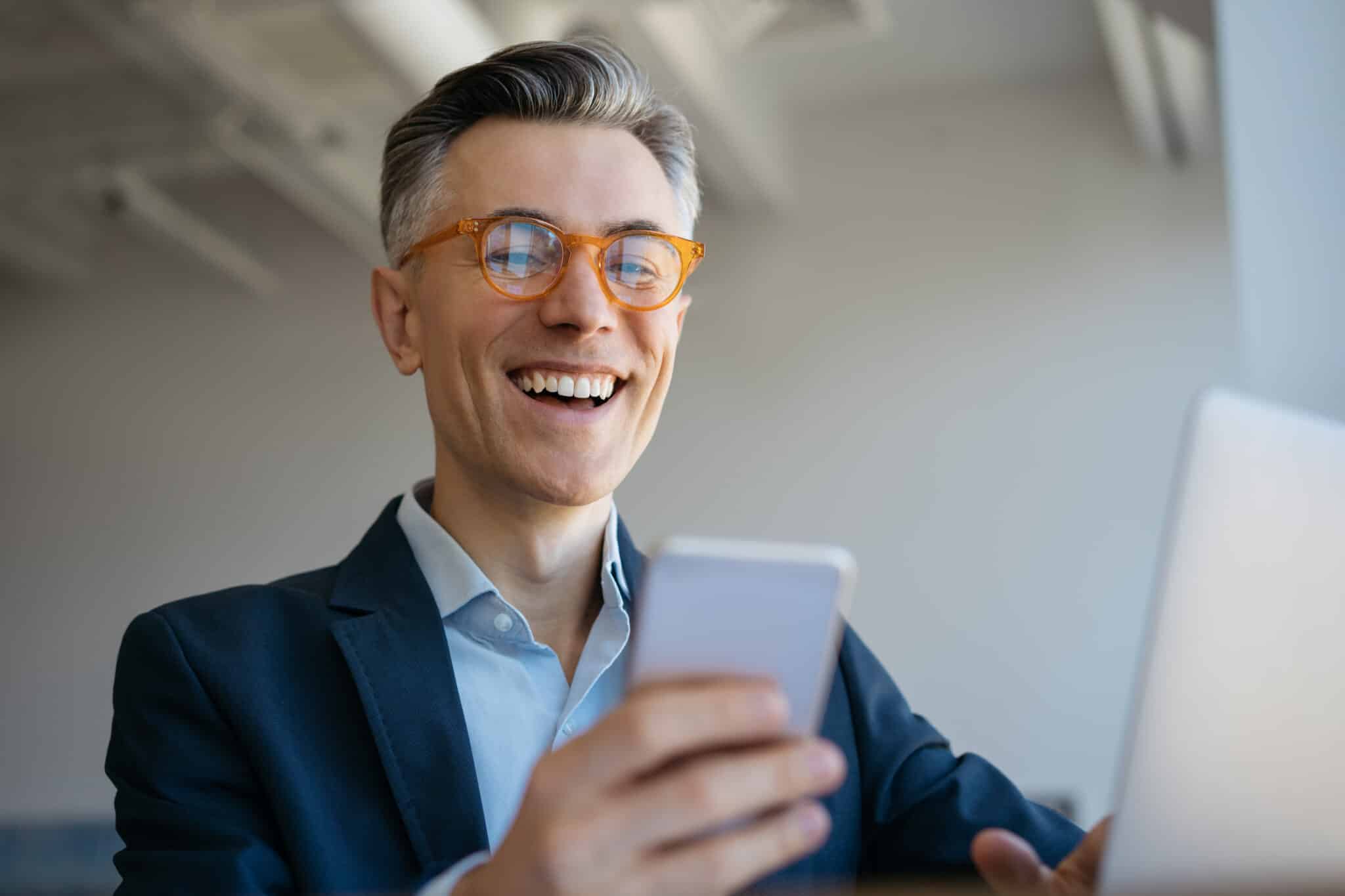 Smiling mature man using laptop computer, holding mobile phone sitting in modern office, successful business. Portrait of handsome emotional freelancer receive payment, working from home