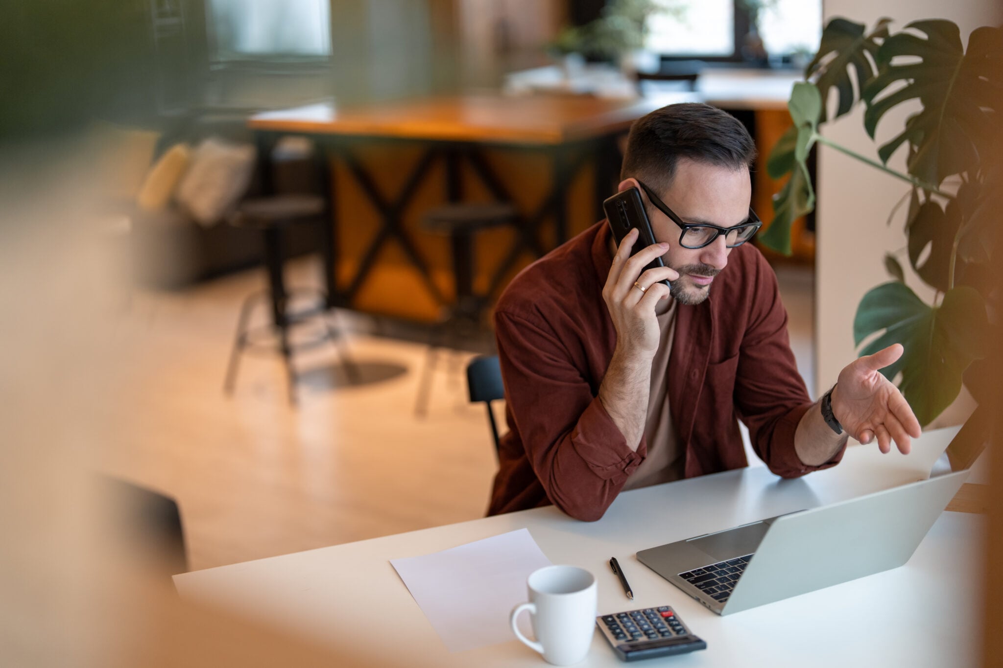A focused man in glasses takes a phone call in a bright café, gesturing as he speaks.