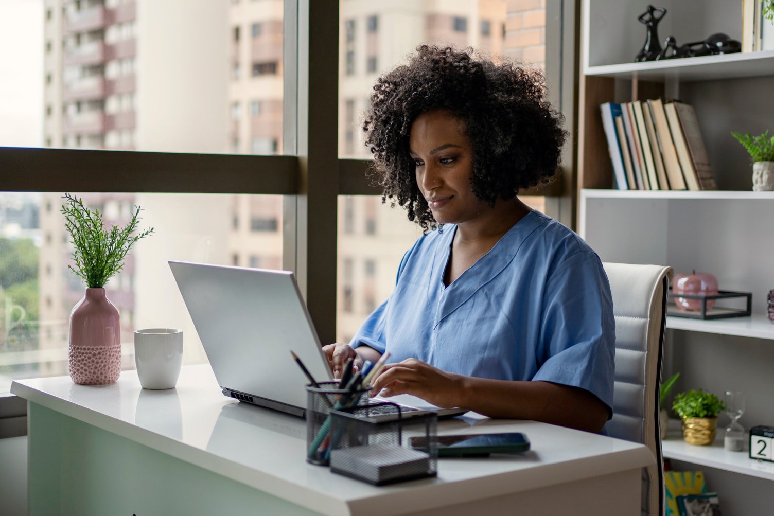 A woman in scrubs sits at a desk, working on a laptop, focused on her task.