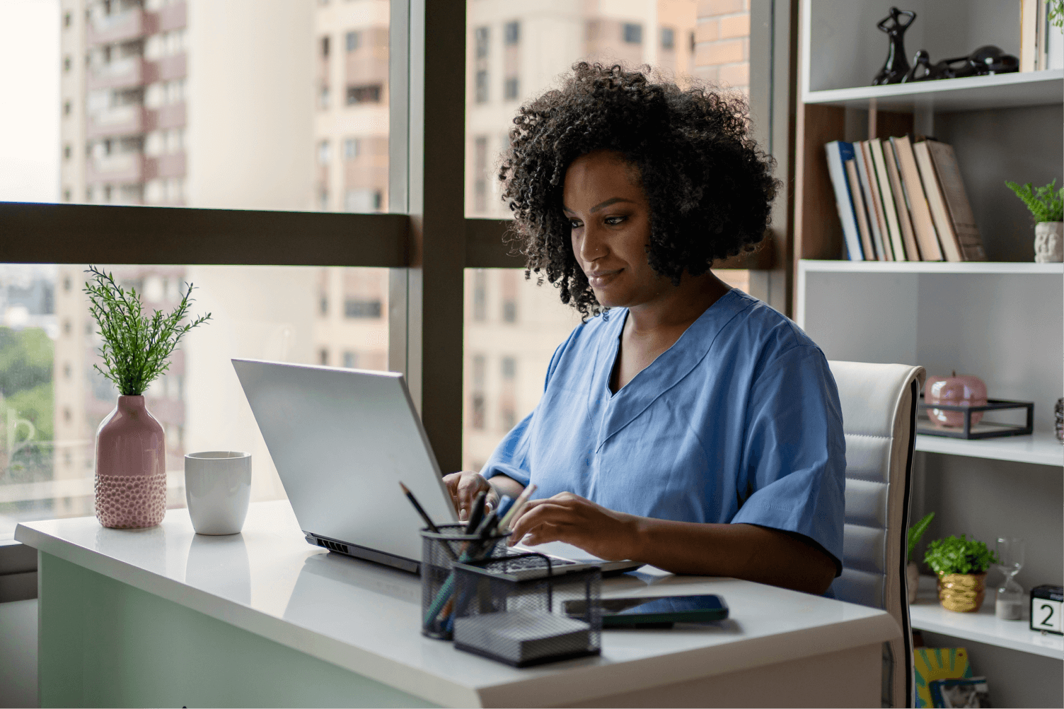 A woman in scrubs sits at a desk, working on a laptop, focused on her task.