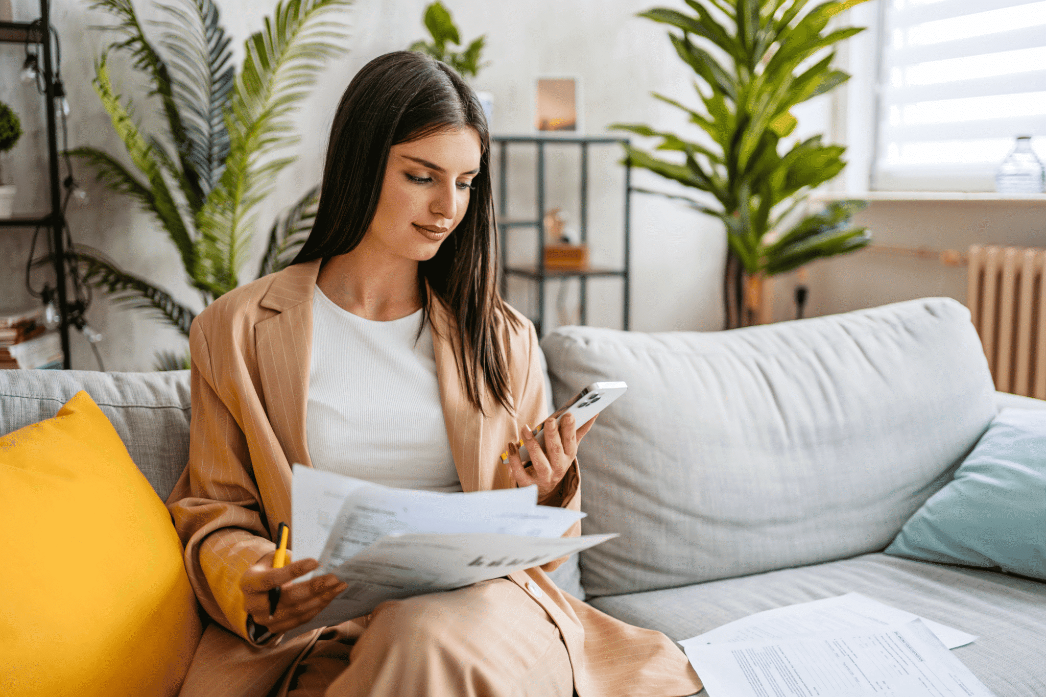 A woman in a phone call on her mobile phone while sitting on a couch and looking at financial documents