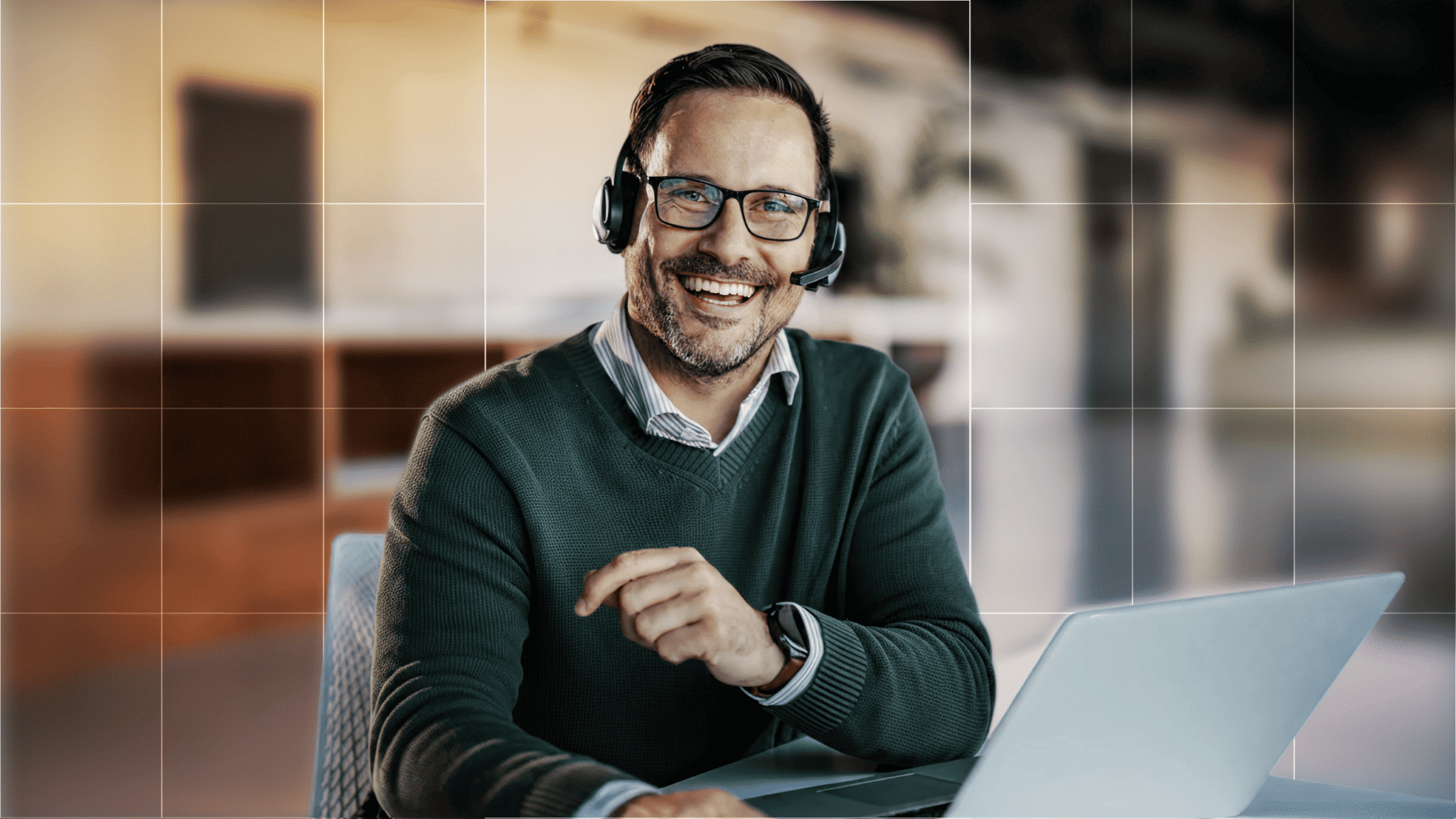 A man wearing glasses and a headset works at a desk with a laptop in front of him.